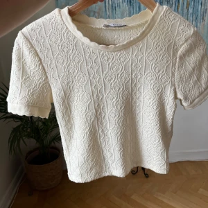 Creamfärgad stickad blus med kort ärm - Supersöt vit stickad blus med korta ärmar och diskret mönsterstickning. Rund halsringning med vågig kant och mjuk struktur. Perfekt för dig som gillar en romantisk och vintageinspirerad stil.