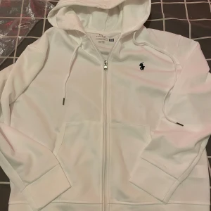 Vit hoodie från Polo Ralph Lauren - Vit hoodie från Polo Ralph Lauren  helt ny med dragkedja och klassisk huva. Tröjan har en svart broderad logga på bröstet, fickor framtill och snörning i huvan. Tillverkad i mjuk bomullsblandning, perfekt för en chill och stilren look.