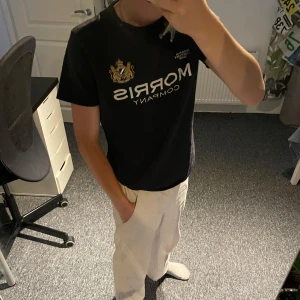 Svart t-shirt från Morris  - Svart t-shirt från Morris Company med stort broderat logotryck och guldfärgat emblem på bröstet. Klassisk rund hals och korta ärmar. Skön passform och stilren design, perfekt för dig som gillar snygga märkes tishor 