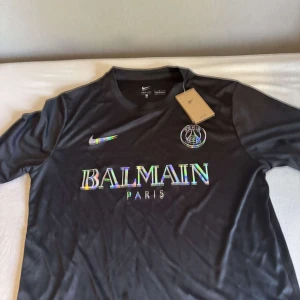 Svart PSG x Balmain fotbollströja Nike - Svart fotbollströja från Nike i samarbete med PSG och Balmain. Tröjan har korta ärmar, rund hals och är tillverkad i Dri-FIT-material som andas. Framsidan har holografiskt Balmain Paris-tryck och PSG-logga. Perfekt för dig som vill sticka ut på planen. Tröjan är helt ny och är i storlek L. Hör av er🦩🦩