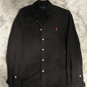 Svart skjorta från Ralph Lauren - Svart långärmad skjorta från Ralph Lauren med klassisk krage och knäppning framtill. Skjortan har en röd broderad logga på bröstet och vita knappar. Snygg och stilren modell i bomull som passar perfekt till jeans eller chinos.