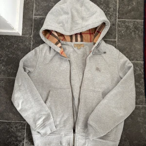 Grå Burberry Zip - Grå Zip hoodie från Burberry med klassiskt rutigt foder i huvan. Jackan har dragkedja framtill, två fickor och ribbade muddar. Perfekt för dig som gillar stilren design med en touch av lyx från det ikoniska brittiska märket.