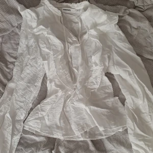 Figursydd Vit volangblus  - Supersöt vit blus från Kappahl med volangdetaljer längs bröstet och axlarna. Långärmad och tillverkad i ett lätt och luftigt bomullstyg. Perfekt för dig som gillar romantisk och feminin stil. Blusen har knytband vid halsen för extra detalj. Figursydd.