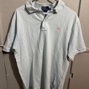 Ljusblå Polo Ralph Lauren t-shirt XL - Snygg ljusblå t-shirt från Polo Ralph Lauren i klassisk pikémodell med krage och knappar. Broderad rosa logga på bröstet. Skön passform och tillverkad i bomull, perfekt för en clean och stilren look.