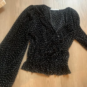 Prickig omlottblus från Pull&Bear - Svart omlottblus från Pull&Bear med vita prickar, långa ärmar och knytband i midjan. Blusen har v-ringning, volangkant nertill och är gjord i ett glansigt, lätt plisserat material som ger en snygg struktur.