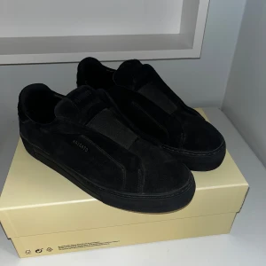 Svarta sneakers från Axel Arigato mocka - Stilrena svarta sneakers från Axel Arigato i mjuk mocka med elastisk panel över vristen istället för klassisk snörning. Snygg minimalistisk design med diskret logga i guld på sidan och helsvart platt sula. Perfekta för dig som gillar cleana och moderna skor. pris gpr att diskutera 