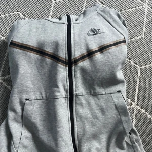 Grå zip-hoodie från Nike med huva - Säljer min Nike tech hoodie i storlek S, tröjan är knappt använd (3 gånger) eftersom den är för liten för mig. Det är en hoodie med ett väldigt skönt material, den köptes i Nikes butik på Emporia så den är självklart äkta. Tveka inte att ställa frågor och pris kan diskuteras
