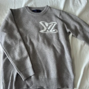 Grå Louis Vuitton stickad tröja - Snygg grå stickad tröja från Louis Vuitton med stort vitt LV-logo på bröstet. Tröjan har rund halsringning, ribbade muddar och är långärmad. Perfekt för dig som gillar exklusiva och stilrena plagg med en lyxig touch.