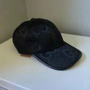 Svart keps med GG-mönster - Snygg svart keps med det klassiska GG-mönstret över hela kepsen. Kepsen har böjd skärm, justerbar rem bak med gulddetalj och är tillverkad i ett slitstarkt tyg med exklusiv känsla. Perfekt accessoar för att lyfta din outfit.