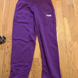 Lila mjukisbyxor från Puma - Snygga lila mjukisbyxor från Puma med vit logga på låret. Byxorna har en bekväm resår i midjan och är gjorda i mjukt bomullsmaterial. Perfekta för chill eller träning.