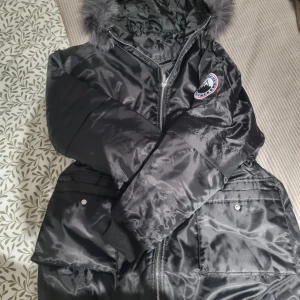 Svart pufferjacka med fuskpäls - Snygg svart pufferjacka med glansig finish och huva med fuskpäls. Jackan har dragkedja framtill, ribbade muddar och stora fickor med lock. Märkespatch på bröstet ger en cool detalj. Perfekt för kalla dagar när du vill hålla stilen.