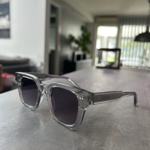 Grå transparenta solglasögon CHIMI - Snygga solglasögon från CHIMI med transparent grå båge och fyrkantig form. Glasen är mörka och ger en cool vibe. Kommer i originalfodral med CHIMI-logga. Perfekta för dig som vill sticka ut med stilrena accessoarer.