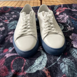 Vita sneakers från Axel Arigato - Snygga vita sneakers från Axel Arigato med stilren design och blå sula. Skorna har rund tå, vita snören och är tillverkade i skinn. Perfekta för dig som gillar minimalistisk och clean stil.