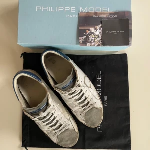 Philippe Model vita och grå sneakers Strlk 45 - Snygga sneakers från Philippe Model i vitt skinn med grå mockadetaljer på tån och blå hälkappa. Klassisk låg modell med snörning och rund tå. Perfekta för dig som gillar en stilren look.