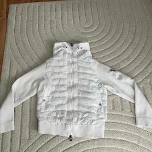 Vit Moncler pufferjacka med huva - Snygg vit pufferjacka från Moncler med quiltad framsida och ribbade ärmar. Jackan har dragkedja, två fickor fram och en huva. Perfekt för dig som vill ha en stilren och sportig look med exklusiv känsla.