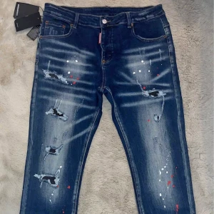 Dsquared2 blå distressed jeans - Snygga blå jeans från Dsquared2 med slitna detaljer, färgstänk i vitt och rött samt tydlig logga bak. Jeansen har klassisk femficksdesign, normal passform och är tillverkade i bomull. Perfekta för dig som gillar en edgy streetstil.
