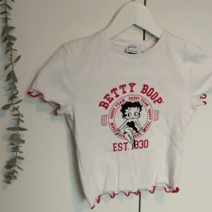 Bershka Betty Boop Cropped T shirt Baby Tee - En vit cropped baby tee / t shirt med svarta och röda detaljer. Betty Boop figur. Med röd lettuce hem. Från Bershka. Storlek: S. Använd ett par antal gånger, i bra/fint skick. 