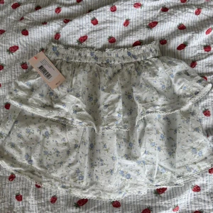 Blommig volangkjol från Nelly - Supersöt kort volangkjol från Nelly i vitt med ljusblått blommönster. Kjolen har elastisk midja, spetsdetaljer och dubbla volanger för en romantisk vibe. Perfekt för sommarens alla tillfällen!