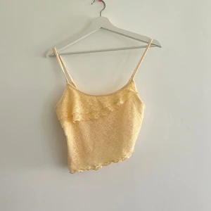 Gul spetsig linnetopp med volang - Söt ljusgul linnetopp med tunna axelband och volangdetalj framtill. Toppen är croppad och har ett fint spetsmönster över hela tyget samt vågig kant nedtill. Perfekt för varma dagar och ger en romantisk vibe. Aldrig använd. 