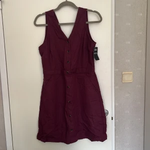 Vinröd/Mörk lila ärmlös klänning från Kühl - Vinröd kortklänning från Kühl i bomull/linne-mix. Klänningen är ärmlös med v-ringning fram och snygg omlott-detalj i ryggen. Knappar framtill och dragkedja bak. Perfekt för dig som gillar en chill men ändå stilren look för sommaren! Storlek: Small/Medium, kan passa både och