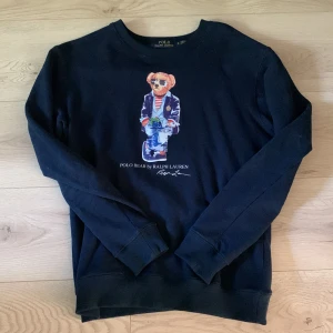 Svart sweatshirt Polo Bear Ralph Lauren - Svart sweatshirt från Polo Ralph Lauren med ikoniska Polo Bear-trycket framtill. Tröjan har rund hals, långa ärmar och ribbade muddar. Perfekt för dig som gillar klassisk streetstyle med en twist. Materialet är mjuk bomull som känns skönt mot huden.