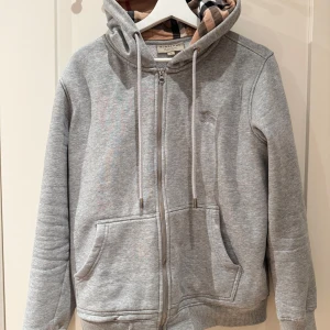 Grå hoodie från Burberry med dragkedja - Snygg grå hoodie från Burberry med klassiskt rutigt foder i huvan. Tröjan har dragkedja, snörning i huvan och två stora fickor framtill. Perfekt för dig som gillar stilren design med en touch av lyx.