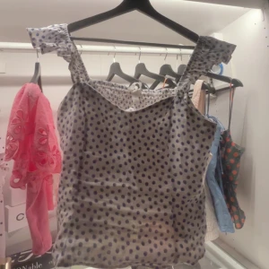 Prickig vit topp med volangaxelband - Supersöt vit topp med blå prickar och volangiga axelband. Sitter figurnära med en lätt v-ringning och ärmlös design. Perfekt för sommarens alla tillfällen och ger en lekfull vibe till din outfit.