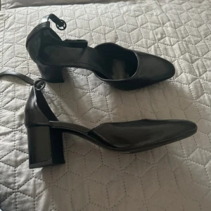 Svarta pumps med blockklack och rem - Stilrena svarta pumps i skinnimitation med fyrkantig tå och justerbar ankelrem. Skorna har en stabil blockklack på ca 6 cm som ger extra höjd och komfort. Perfekta för dig som gillar en klassisk men modern look.