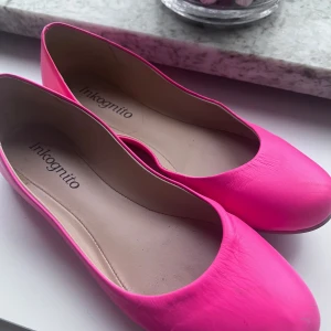 Neonrosa ballerinaskor från Inkognito - Snygga ballerinaskor i neonrosa från Inkognito. Skorna har rund tå och platt sula, tillverkade i syntetmaterial. Perfekta för dig som vill sticka ut med färg och enkel stil. Storlek 36.