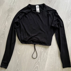 Svart croppad långärmad tröja Gymshark - Svart långärmad tröja från Gymshark med croppad passform och snörning i midjan. Tillverkad i mjukt och stretchigt material som passar perfekt till träning. Diskret logga på ryggen och enkel, clean look.