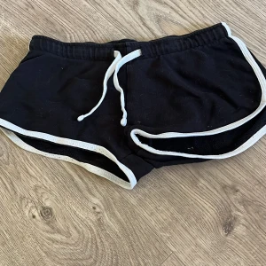 Svarta shorts med vit kant från FASBTER - Svarta sportiga shorts från FASBTER i storlek XS. De har vit kant runtom och vit snörning i midjan för en cool kontrast. Perfekta för sommardagar eller träning, med en loose passform och mjukt bomullsmaterial som känns skönt mot huden.