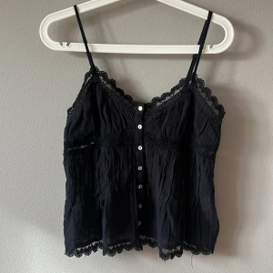 Svart spetslinnetopp från Zara - Supersöt svart linnetopp från Zara med tunna axelband, knappar framtill och fina spetsdetaljer längs kanterna. Toppen är luftig och har en lätt rynkad struktur som ger en avslappnad känsla. Perfekt att styla med jeans eller kjol för en trendig look.