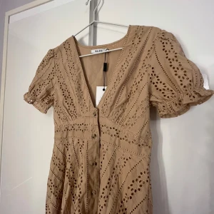 Oanvänd beige kort klänning från NA-KD - Supersöt beige kort klänning från NA-KD med broderade hål, puffärmar och v-ringning. Klänningen har knappar framtill och är tillverkad i bomull. Perfekt för sommarens alla tillfällen och ger en fräsch och trendig look. Prislappen sitter kvar!