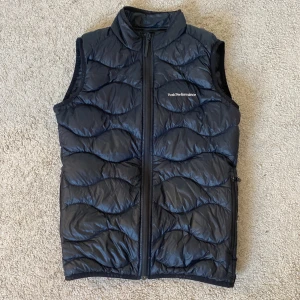 Svart dunväst från Peak Performance - Säljer en svart dunväst från Peak Performance med vågformat quiltat mönster. Västen har hög krage, dragkedja framtill och diskret logga på bröstet. Perfekt för lager-på-lager och enkel att matcha med andra plagg.