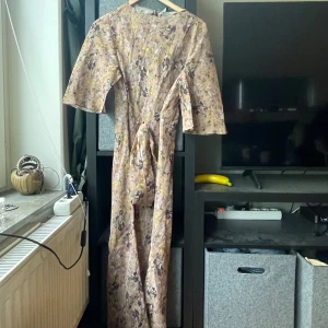 Blommig långklänning från & Other Stories - Superfin långklänning från & Other Stories med blommigt mönster i ljusa toner av beige, lila och gult. Klänningen har trekvartsärm, knytband i midjan och dragkedja bak. Tillhörande underklänning i beige medföljer. Perfekt för sommarens alla tillfällen!