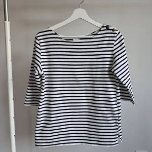 Randig tröja med knappar från Oysho M 38 - Säljer en vit och svart randig tröja från Oysho med trekvartsärm och två snygga guldknappar med ankare på axeln. Tröjan är i mjuk bomull och har en klassisk båtringad hals. Perfekt för en chill och stilren look.