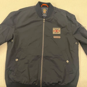 Svart bomberjacka från Morris R Club - Snygg svart bomberjacka från Morris R Club med Union Jack-patch och coola detaljer. Jackan har dragkedja framtill, fickor med tryckknappar och en extra ficka med dragkedja på ärmen. Ribbstickade muddar och klassisk bomberstil. Perfekt för dig som gillar streetwear. (Aldrig någonsin använd) 