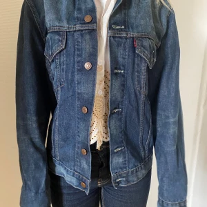  jeansjacka från Levi's - Säljer en klassisk blå jeansjacka från Levi's med knappar framtill och två bröstfickor med lock. Jackan har en tidlös design och är perfekt att slänga över en t-shirt eller blus. Ikonisk röd Levi's-tagg på bröstfickan.