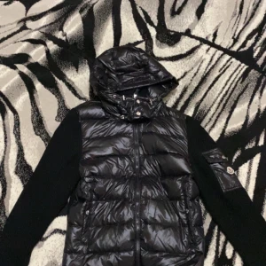 Svart pufferjacka med huva från Moncler - Säljer en svart pufferjacka från Moncler med glansig framsida, stickade ärmar och huva. Jackan har dragkedja, tryckknappar och en ficka med Moncler-logga på ärmen. Perfekt för kyliga dagar.KVITTO FINNS KOM DM ❗️