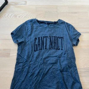 Blå t-shirt från GANT med tryck - Säljer en blå t-shirt från GANT med stort svart tryck 'GANT NHCT' på bröstet. T-shirten har rund halsringning och korta ärmar. Perfekt till jeans eller shorts för en avslappnad stil.