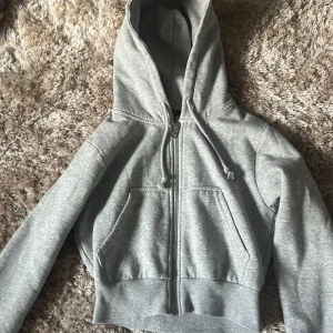 Grå hoodie från Monki - Säljer en klassisk kort grå hoodie från Monki i storlek XS. Tröjan har dragkedja framtill, huva med dragsko och två stora fickor. Perfekt för en avslappnad stil och enkel att matcha med det mesta.
