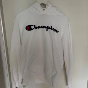 Vit hoodie från Champion - Säljer en klassisk vit hoodie från Champion med broderad logga i blått, rött och svart på bröstet. Tröjan har huva och en stor magficka framtill. Perfekt för en avslappnad och sportig stil.