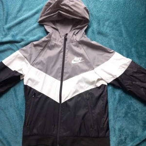 Nike vindjacka med huva i svart, grå och vitt - Nike vindjacka med huva i svart, grå och vitt. Jackan har dragkedja framtill, två sidofickor med dragkedja och stor Nike-logga på både bröstet och ryggen. Perfekt för blåsiga dagar och sportiga outfits.
