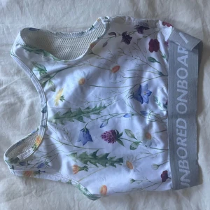 Blommig sport-bh från Eivy - Superfin sport-bh från Eivy med blommigt mönster i flera färger på vit botten. Bred grå resår med texten 'UNBORED ONBOARD'. Perfekt för träning och har mesh på insidan för extra komfort.