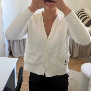 Vit kofta från Polo Ralph Lauren - Säljer en klassisk vit kofta från Polo Ralph Lauren med knappar framtill och två fickor. Tröjan har lång ärm och är stickad i mjukt material. Liten broderad logga i mörkblått nertill på ena sidan.