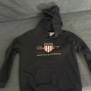 Svart hoodie från Gant - Säljer en svart hoodie från Gant med broderad logga och texten 'American Sportswear' på bröstet. Tröjan har huva och en stor magficka framtill. Perfekt för dig som gillar klassisk och sportig stil. Säljer den pågrund av att jag har växt ifrån den. Använt 3 gånger max