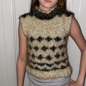 Beige och svart stickad väst med mönster - Mysig stickad väst i beige med svarta detaljer och glittrigt garn. Västen har ett grafiskt mönster och hög krage. Perfekt för lager-på-lager och ger en trendig look.