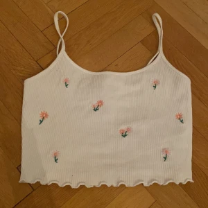 Vit croppad linnetopp med broderade blommor från FB Sister - Superfin vit croppad linnetopp från FB Sister med tunna axelband och små broderade blommor i rosa och grönt. Toppen har ribbad struktur och vågig kant nertill. Perfekt för varma dagar!