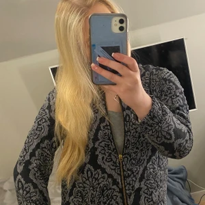 Svart mönstrad jacka med dragkedja - Säljer en svart och grå jacka med stort blommigt mönster och guldfärgad dragkedja framtill. Jackan har lång ärm och är i ett mjukt, stretchigt material. Perfekt för dig som vill sticka ut med en unik stil.