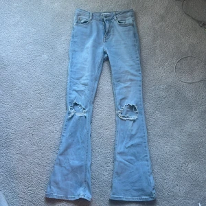 Ljusblå bootcut jeans med slitningar - Säljer ett par ljusblå bootcut jeans med slitna hål på båda knäna. Jeansen har klassisk femficksdesign och normal passform upptill som går ut i vida ben. Perfekta för en avslappnad och trendig look.